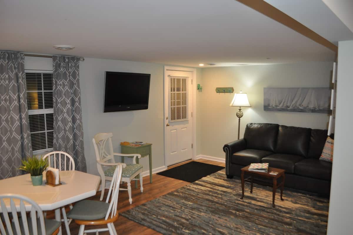 A living room with a black leather sofa, a dining table with chairs, a TV mounted on the wall, and a lamp on a side table.