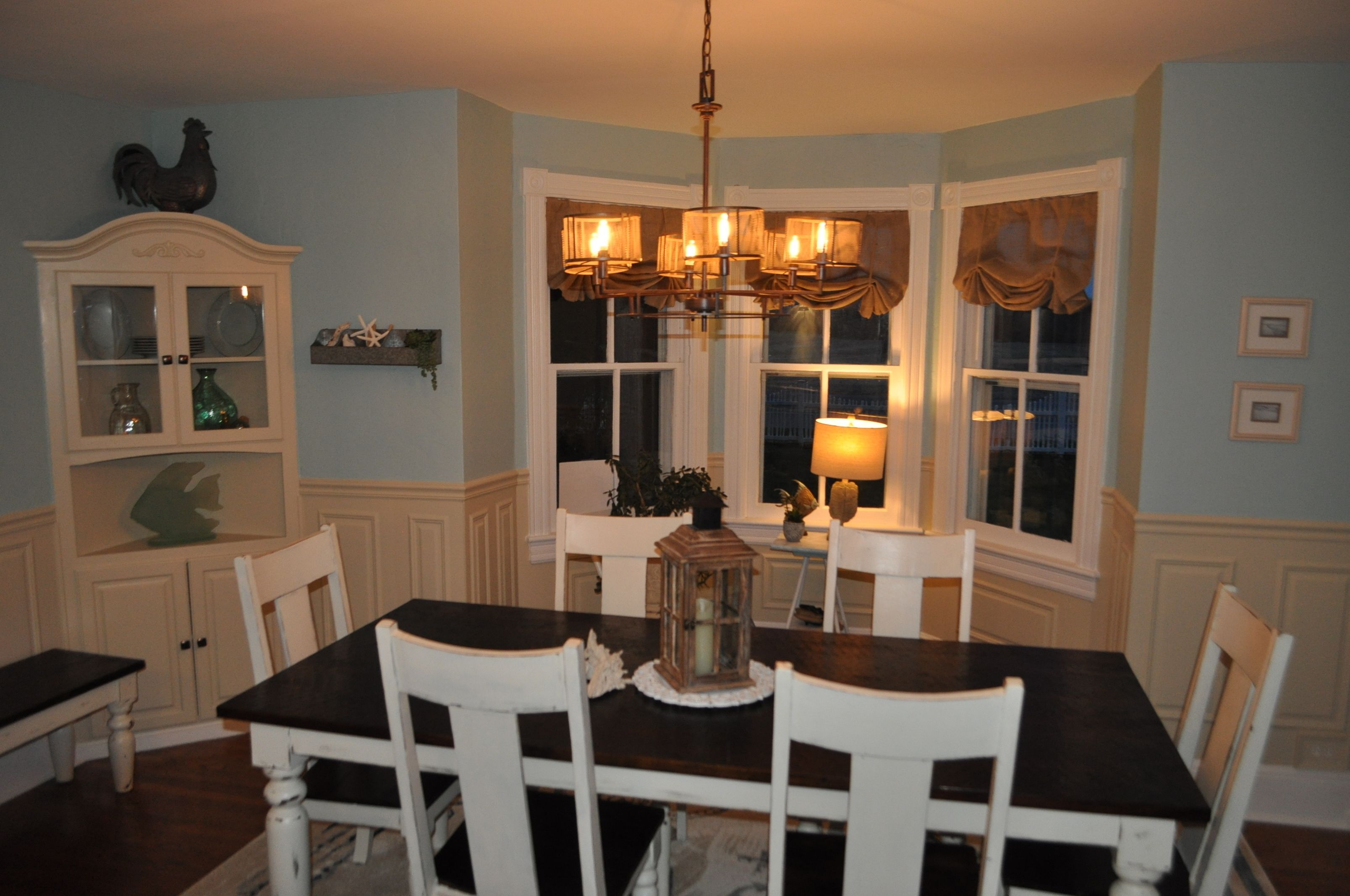 A dining room with a dark wooden table, white chairs, a chandelier, and a window with curtains.