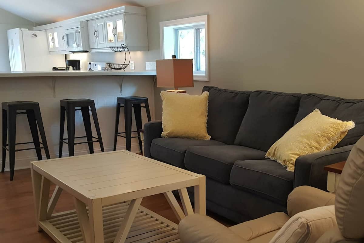 A modern living room with a dark grey sofa, two yellow cushions, a wooden coffee table, and a kitchen area with white cabinets and a black countertop.