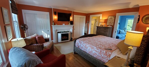 A bedroom with a fireplace, a bed with a patterned bedspread, a red armchair, a dresser, and a television mounted on the wall.