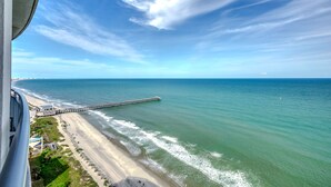 A coastal view with a pier extending into the sea, a beach, and a building with a balcony overlooking the scene. - Fabulous Penthouse, Central MB, 2 Balconies,Watch Sunrise in Your Bed (Myrtle Beach)