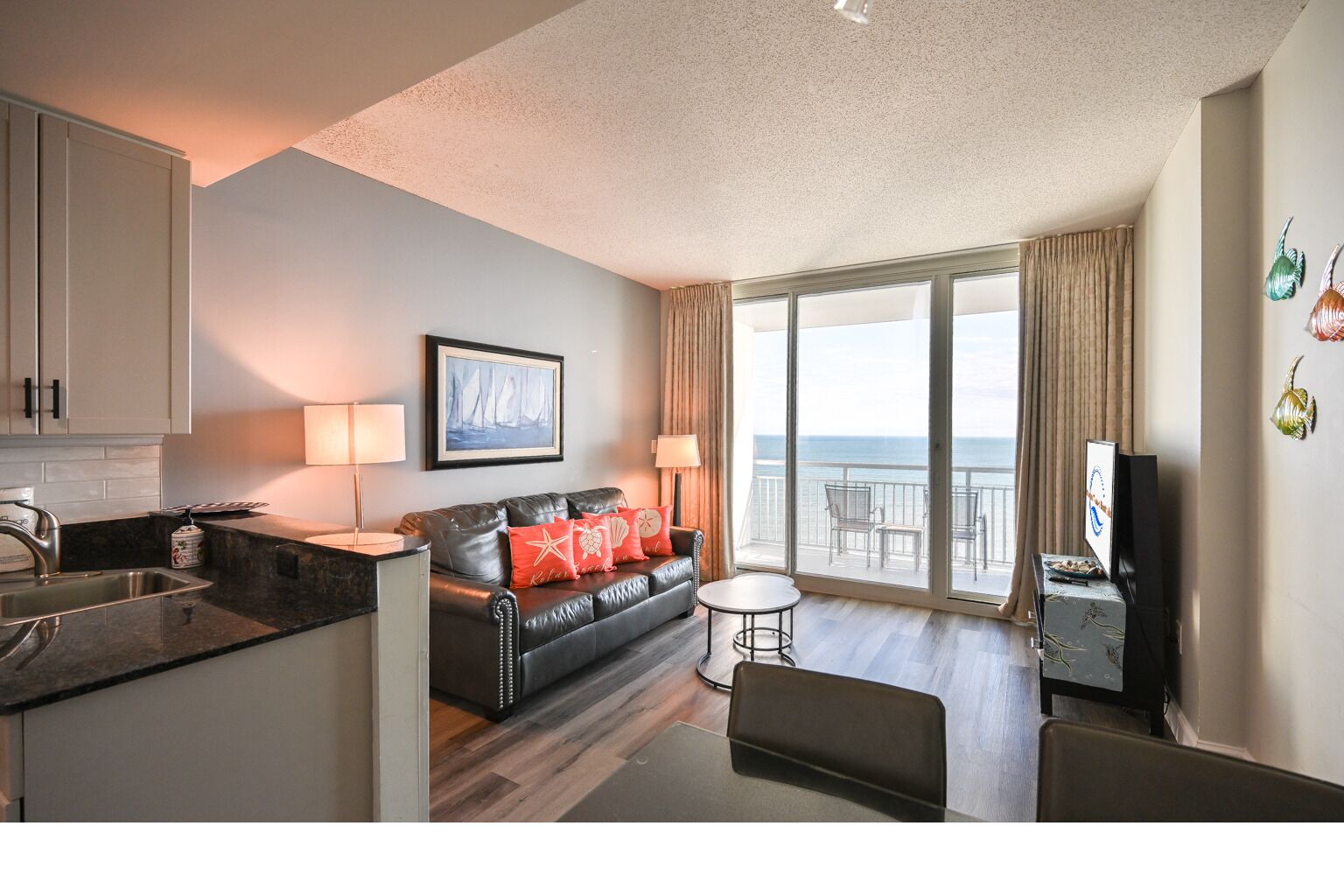 A modern hotel room with a kitchen, a living area with a sofa and a TV, and a balcony with a view of the sea.