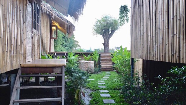 A bamboo structure with a staircase leading to a wooden deck, surrounded by lush greenery and a stone pathway. - Lanna House Lanna Hut Chiangmai (San Kamphaeng)