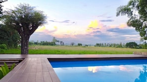 A swimming pool with a wooden deck and a view of a grassy field and trees under a sunset sky. - Lanna House Lanna Hut Chiangmai (San Kamphaeng)