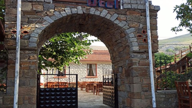 L’entrée d’un hôtel, avec une arche en pierre, une enseigne au-dessus indiquant « Natura Hotel », et une vue sur l’intérieur, où l’on aperçoit des tables et des chaises.