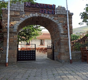 Entrada a un hotel con un arco de piedra, un letrero que dice "Natura Hotel", y vista del interior con mesas y sillas.
