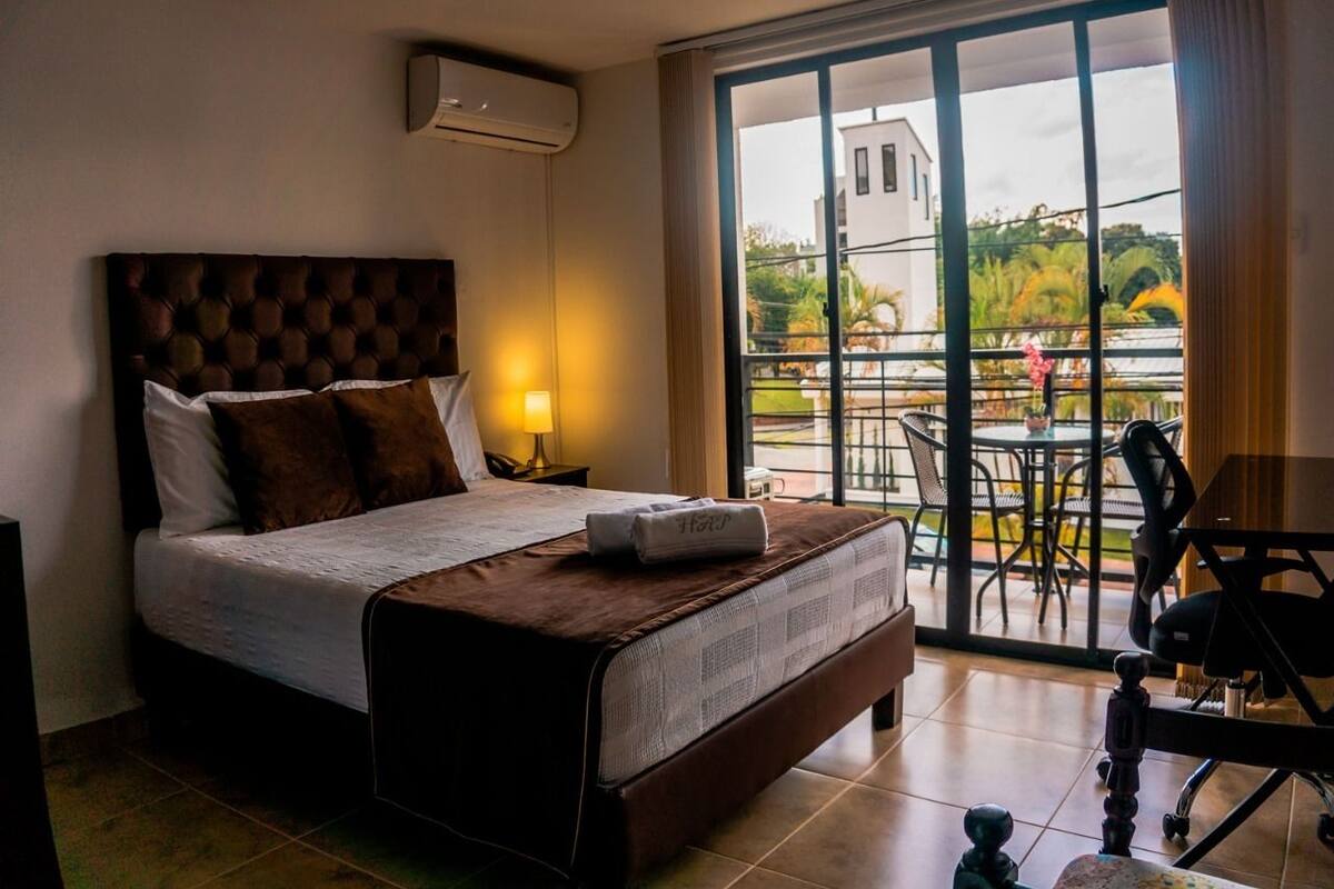 A hotel room with a large bed, a nightstand, a lamp, and a view of the outside balcony.
