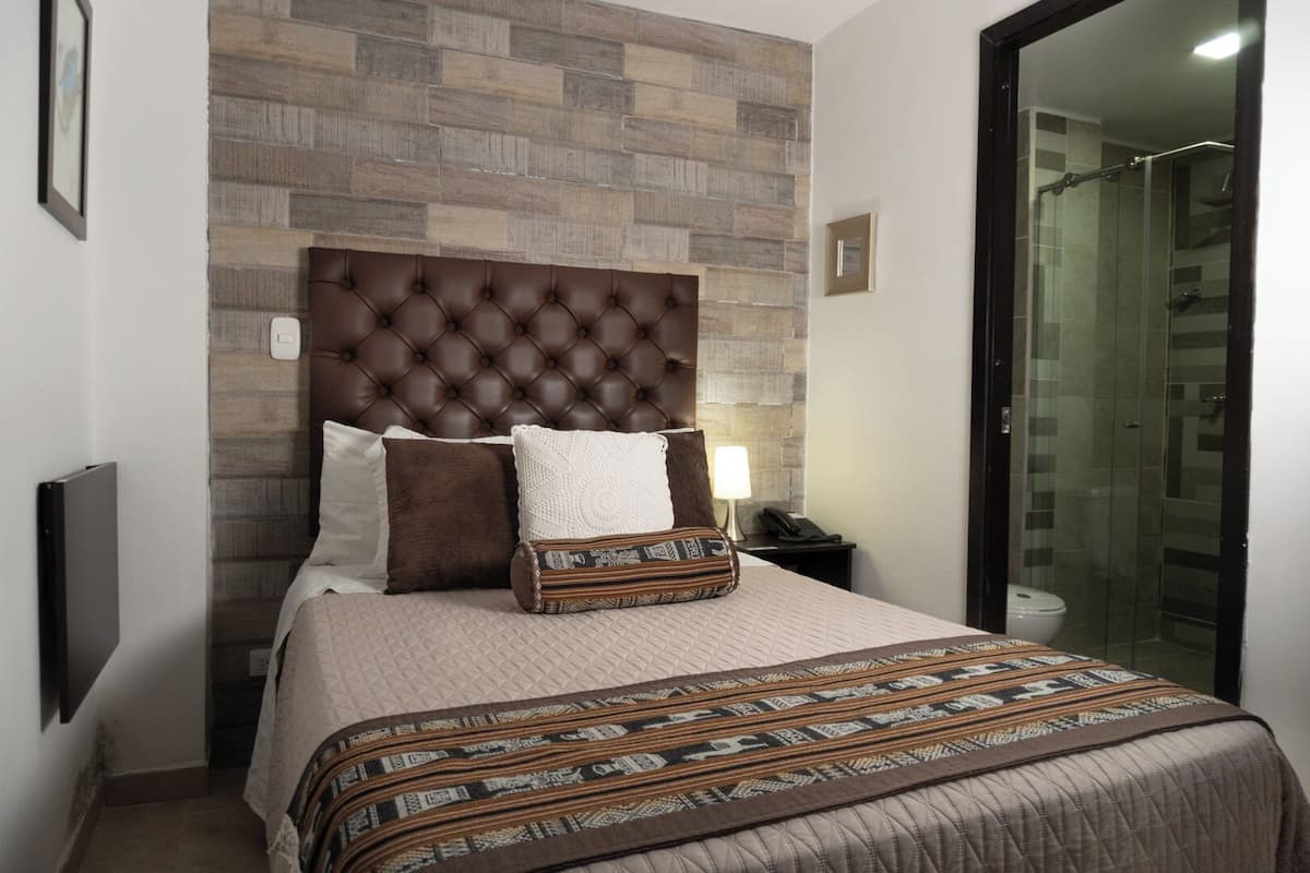 A bedroom with a tufted headboard, a patterned bedspread, a decorative pillow, a bedside lamp, and a glass shower enclosure.