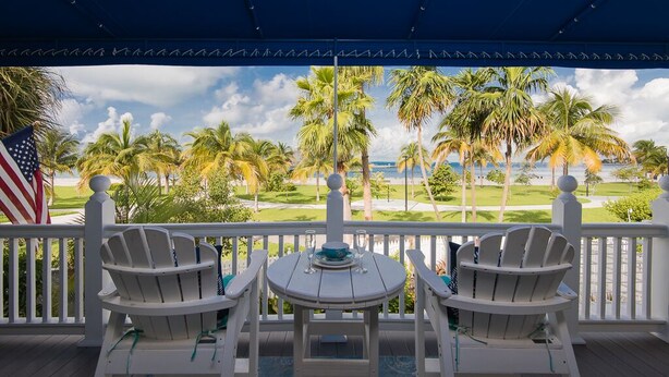 A covered outdoor seating area with two Adirondack chairs and a small table, overlooking a golf course and palm trees.