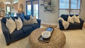 A living room with two blue sofas, a woven coffee table, and a bookshelf with decorative items. - Lagoon Front, Fire Pit, Quiet, Nature ~ Otter Cove (Indian Pass)