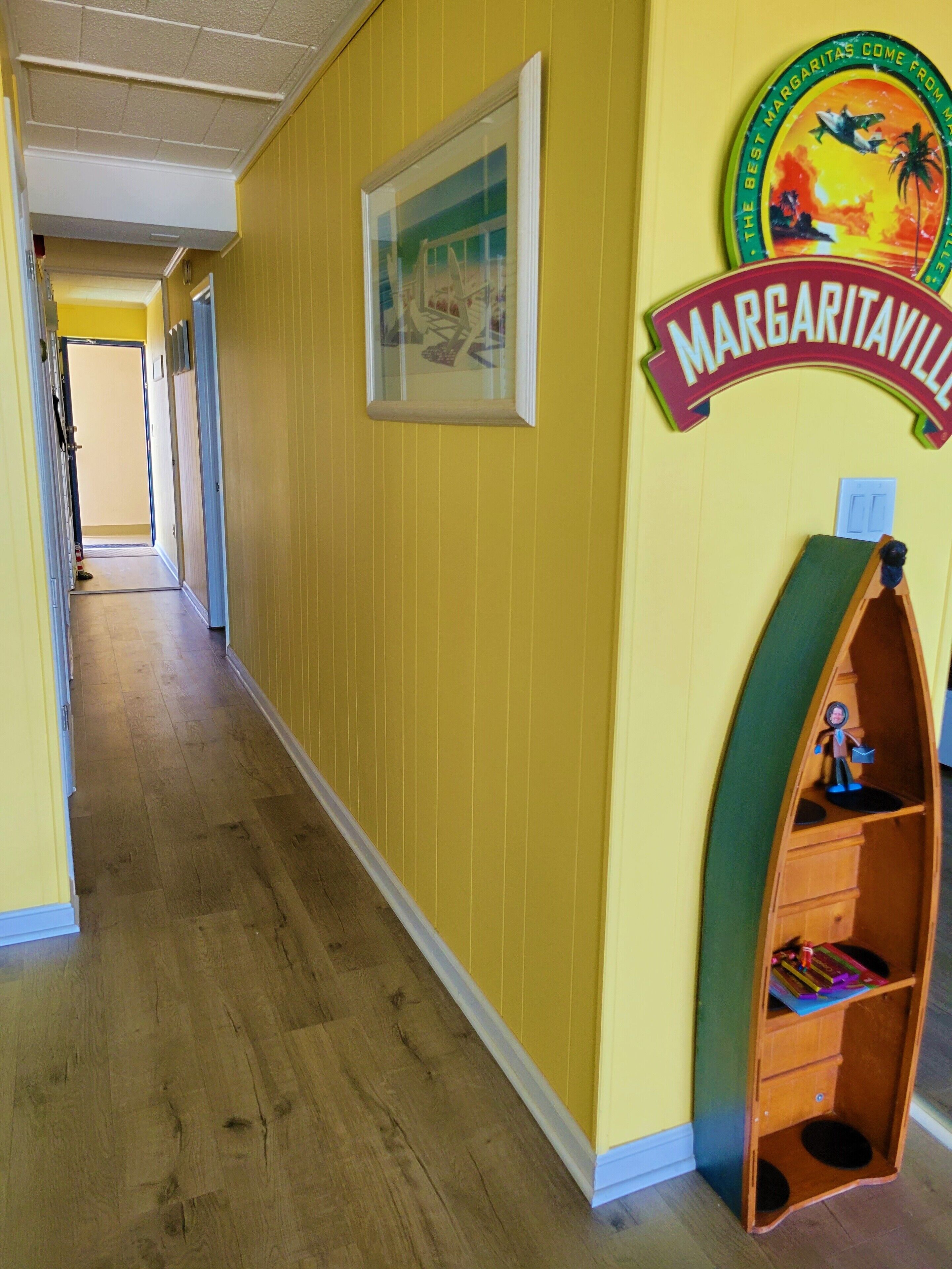 A hallway with yellow walls, a wooden floor, a framed picture, and a shelf with a sign reading "Margaritaville".