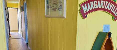 A hallway with yellow walls, a wooden floor, a framed picture, and a shelf with a sign reading "Margaritaville".
