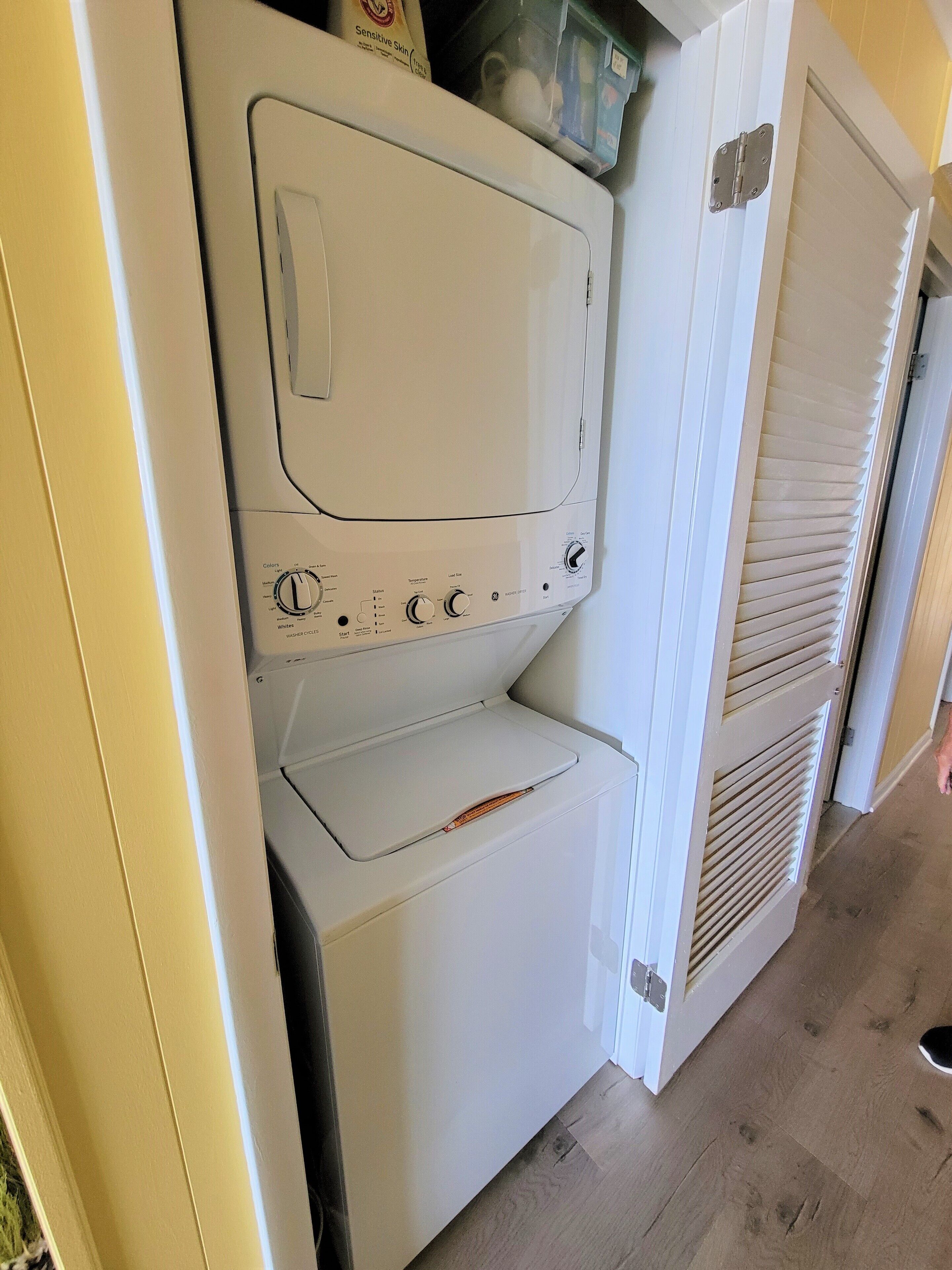 A compact, white laundry room with a built-in washer and dryer, a small shelf with a container, and a door with a window.
