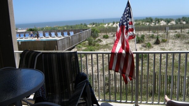 A balcony with a view of the beach, an American flag, and a pool area.