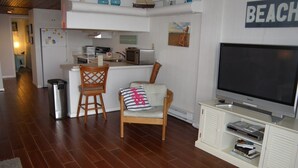 A living room with a TV, a chair, a small table, and a kitchen area in the background.