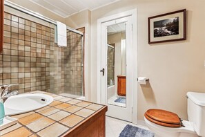 Ein Badezimmer mit gefliestem Waschbecken, einem Holz-Waschtisch, einer Duschkabine aus Glas, einer hölzernen Toilette und einem gerahmten Bild an der Wand.