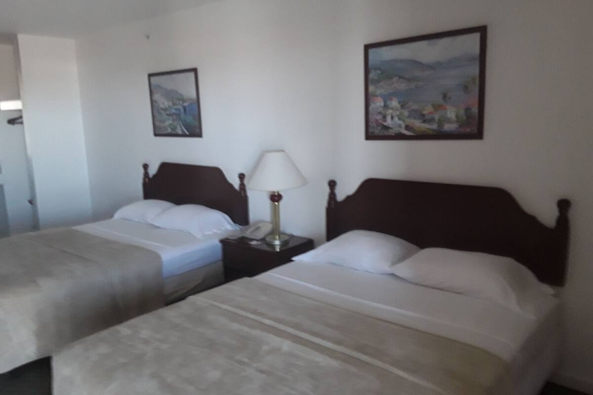 A hotel room with two single beds, a nightstand with a lamp, and two framed pictures on the wall.