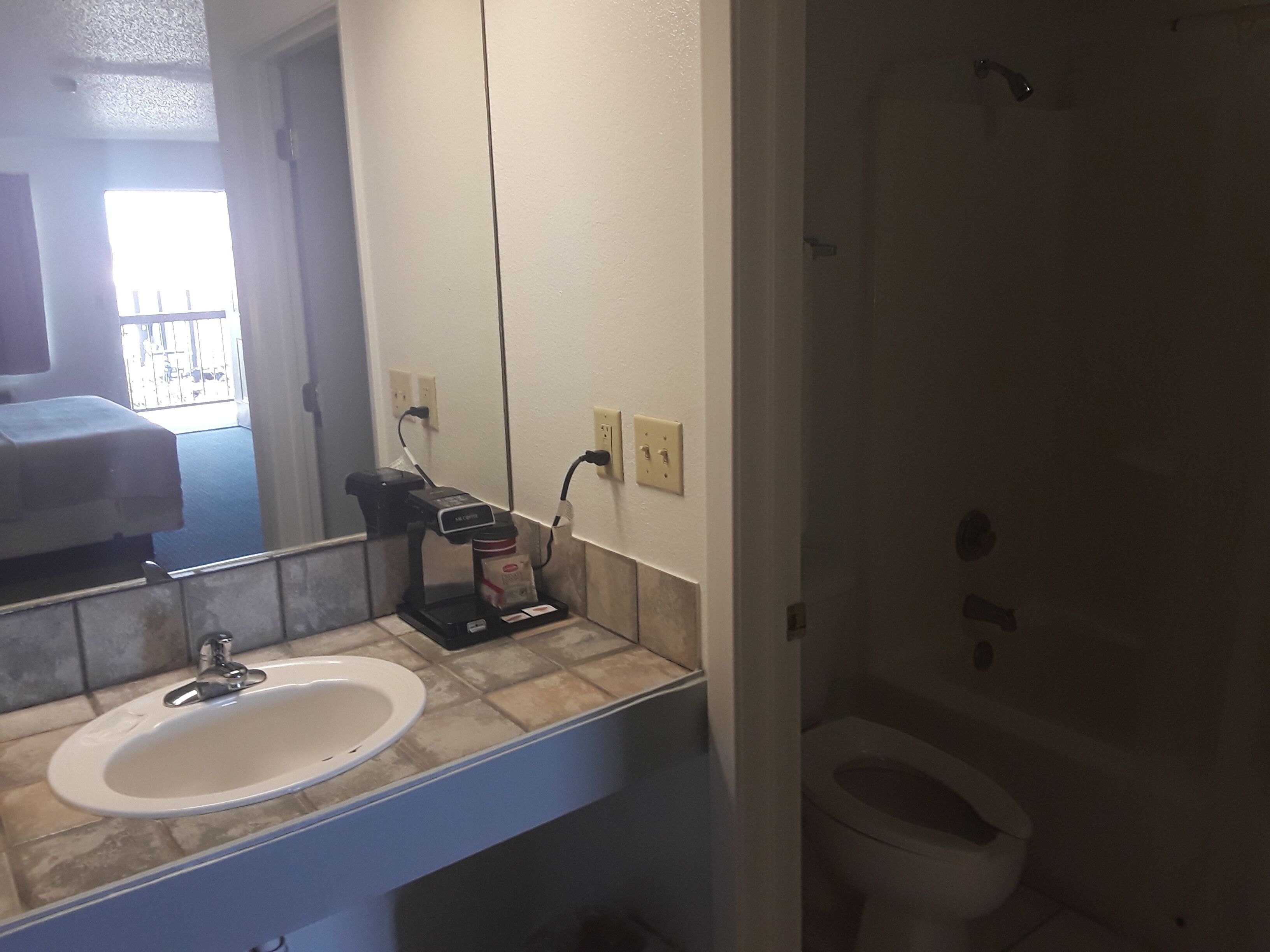 A bathroom with a sink, mirror, and toilet. There is a coffee maker on the counter and a window with a view of the outside.