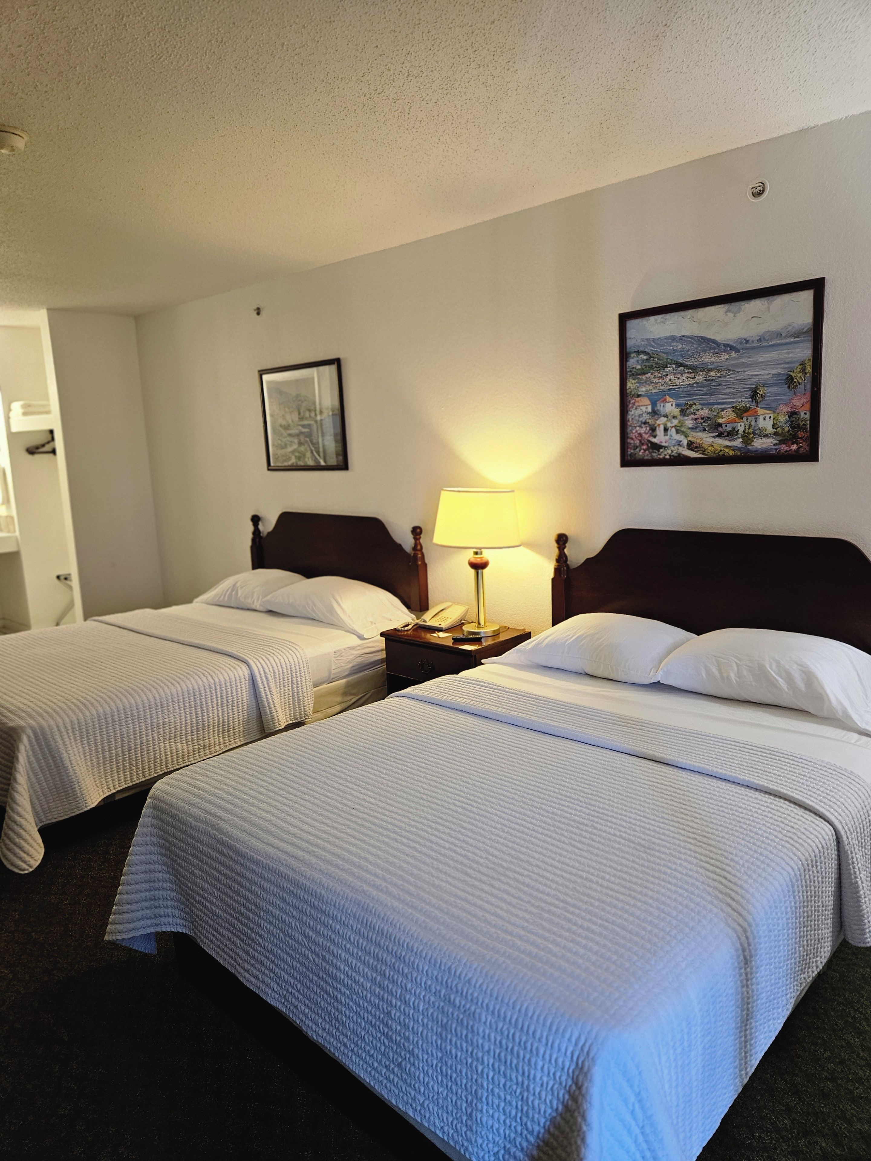 A hotel room with two beds, a nightstand with a lamp, a painting on the wall, and a view of the sea.