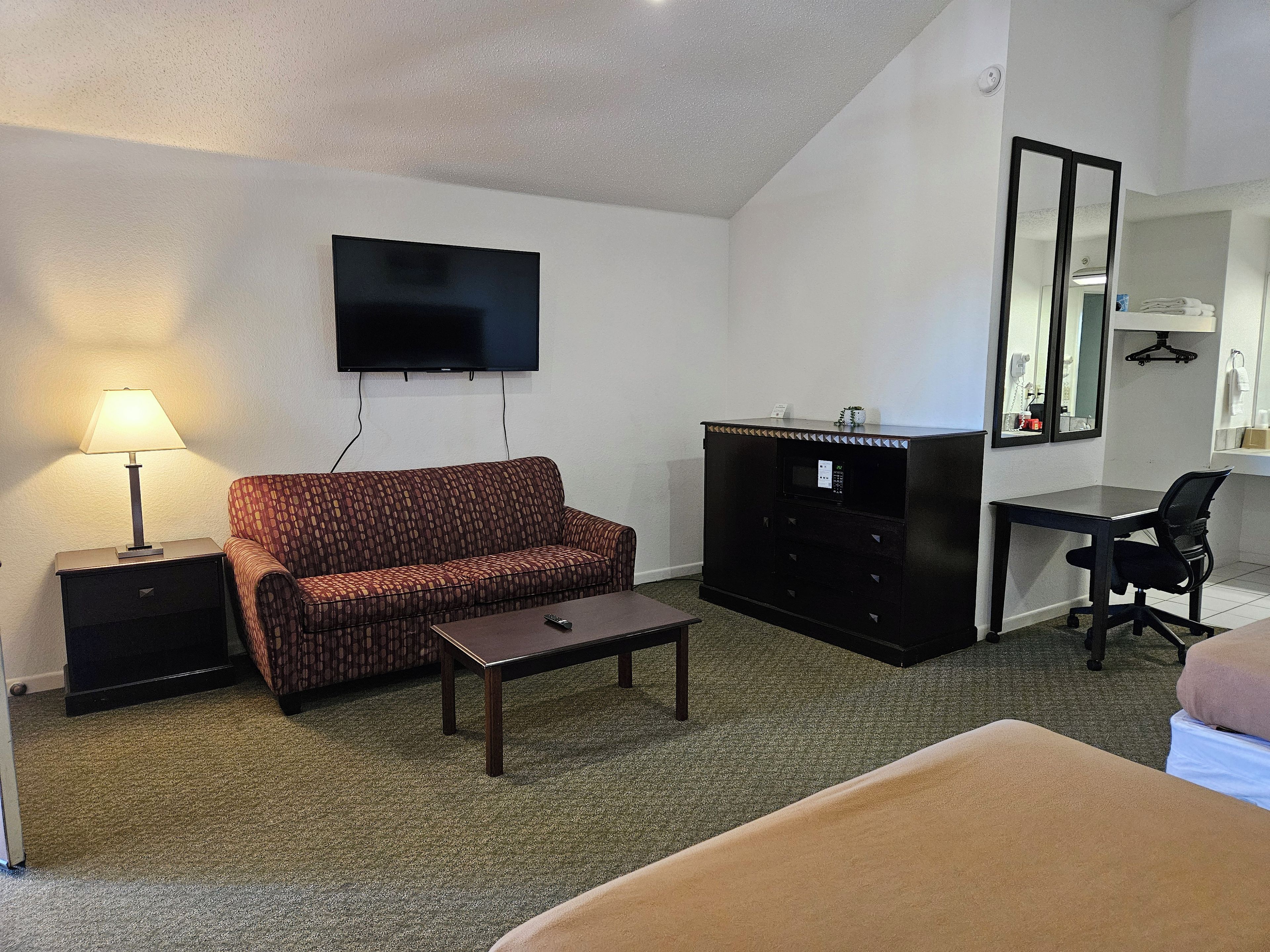 A hotel room with a sofa, a desk, a television, a mirror, and a small cabinet.
