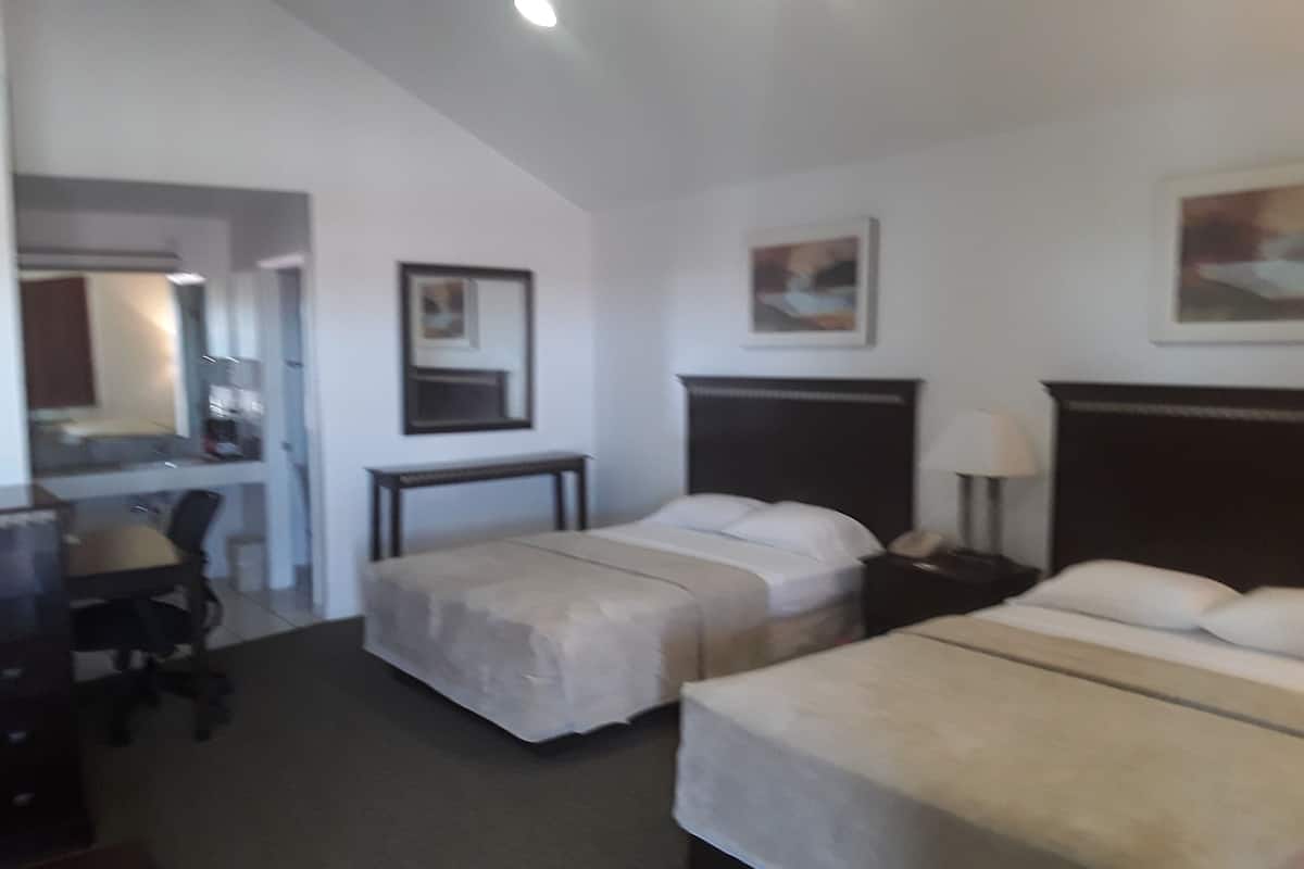 A hotel room with two single beds, a desk with a chair, a small table, and a nightstand with a lamp.