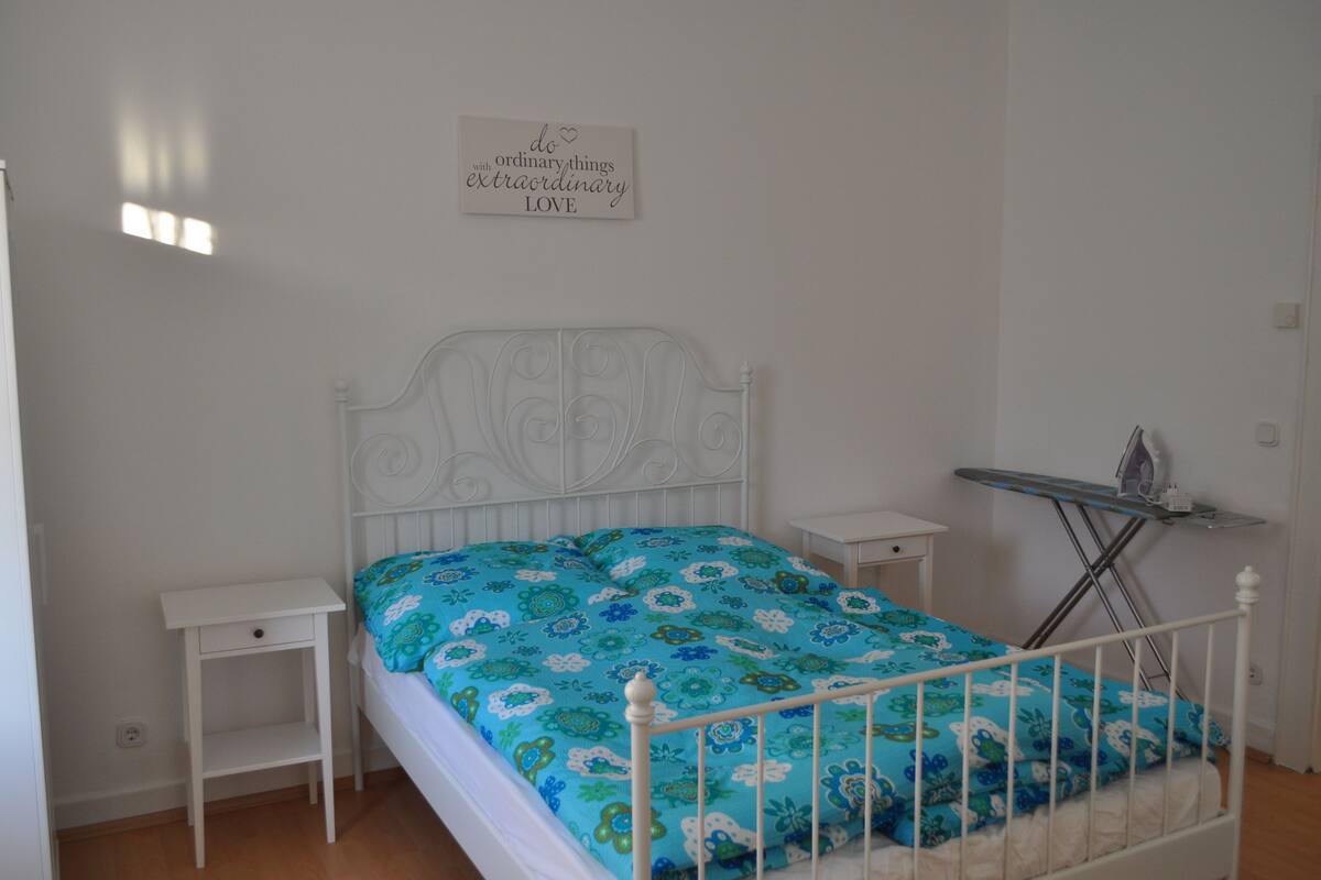 A bedroom with a bed, a nightstand, an ironing board, and a motivational quote on the wall.
