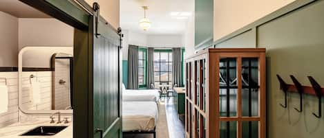 A narrow hotel corridor with a bed, a bathroom, and a seating area.