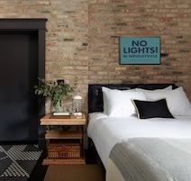 A bedroom with a brick wall, a bed with white bedding, a nightstand with a lamp, and a framed sign that reads "NO LIGHTS! IN WRIGLEYVILLE".
