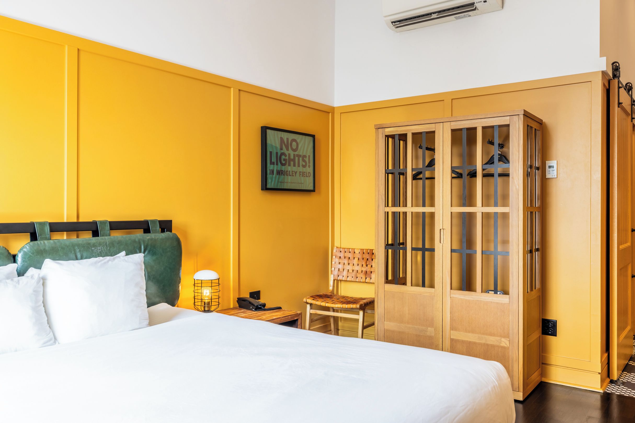 A hotel room with a bed, a wooden wardrobe, a chair, a nightstand, and a wall-mounted air conditioning unit.