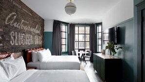 A hotel room with two beds, a brick accent wall, a large window with curtains, a desk with a chair, a TV, and a plant. - The Chicago Hotel Collection - Wrigleyville (Chicago)