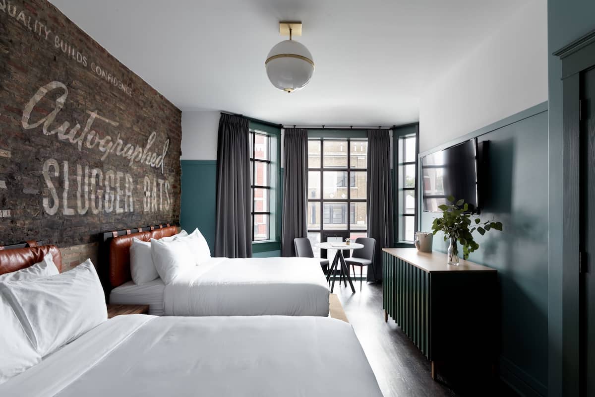 A hotel room with two beds, a brick accent wall, a large window with curtains, a desk with a chair, a TV, and a plant.