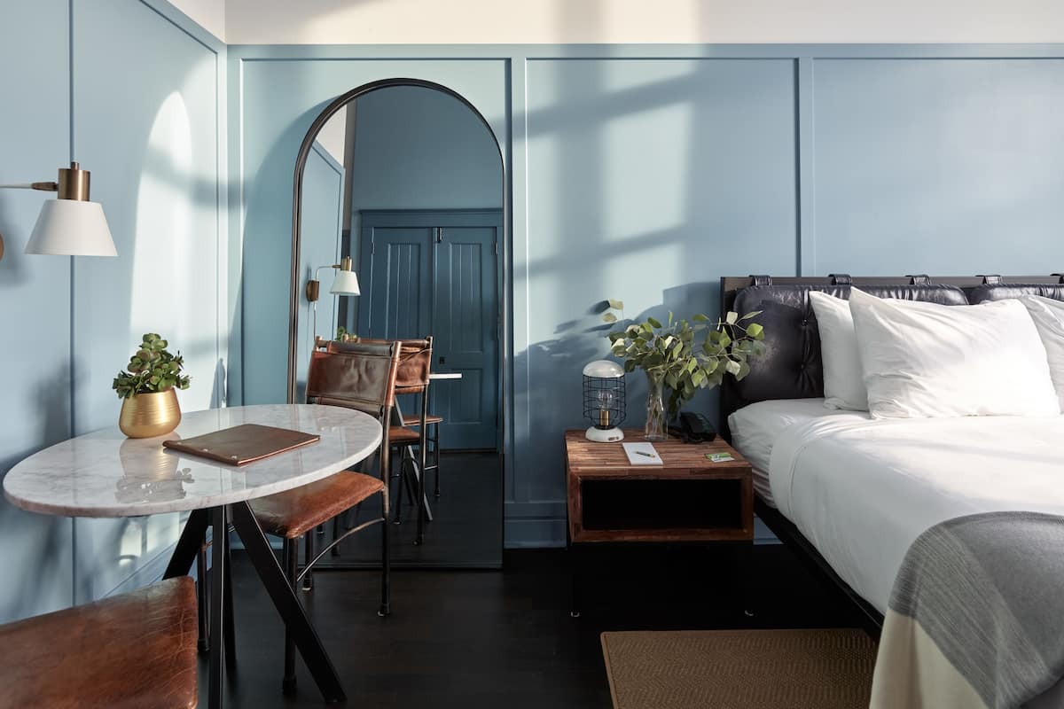 A modern hotel room with a bed, a desk, a chair, and a small table.