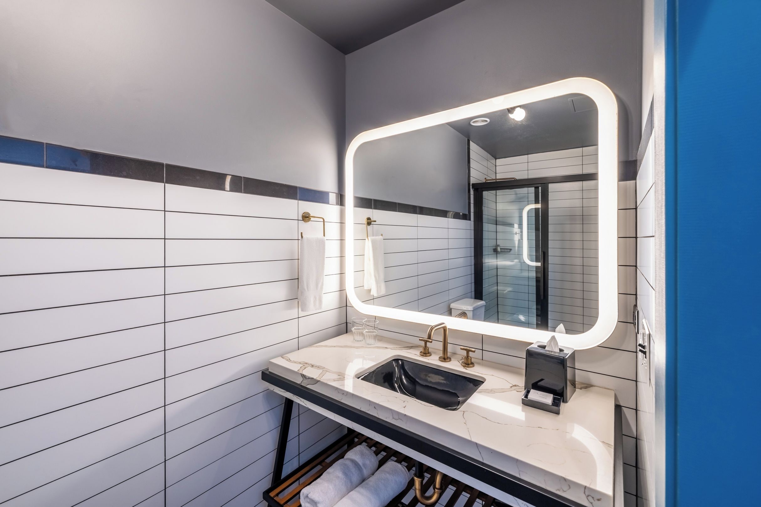 Un baño moderno con un espejo grande, paredes de azulejos blancos, encimera de mármol y un perchero para toallas.