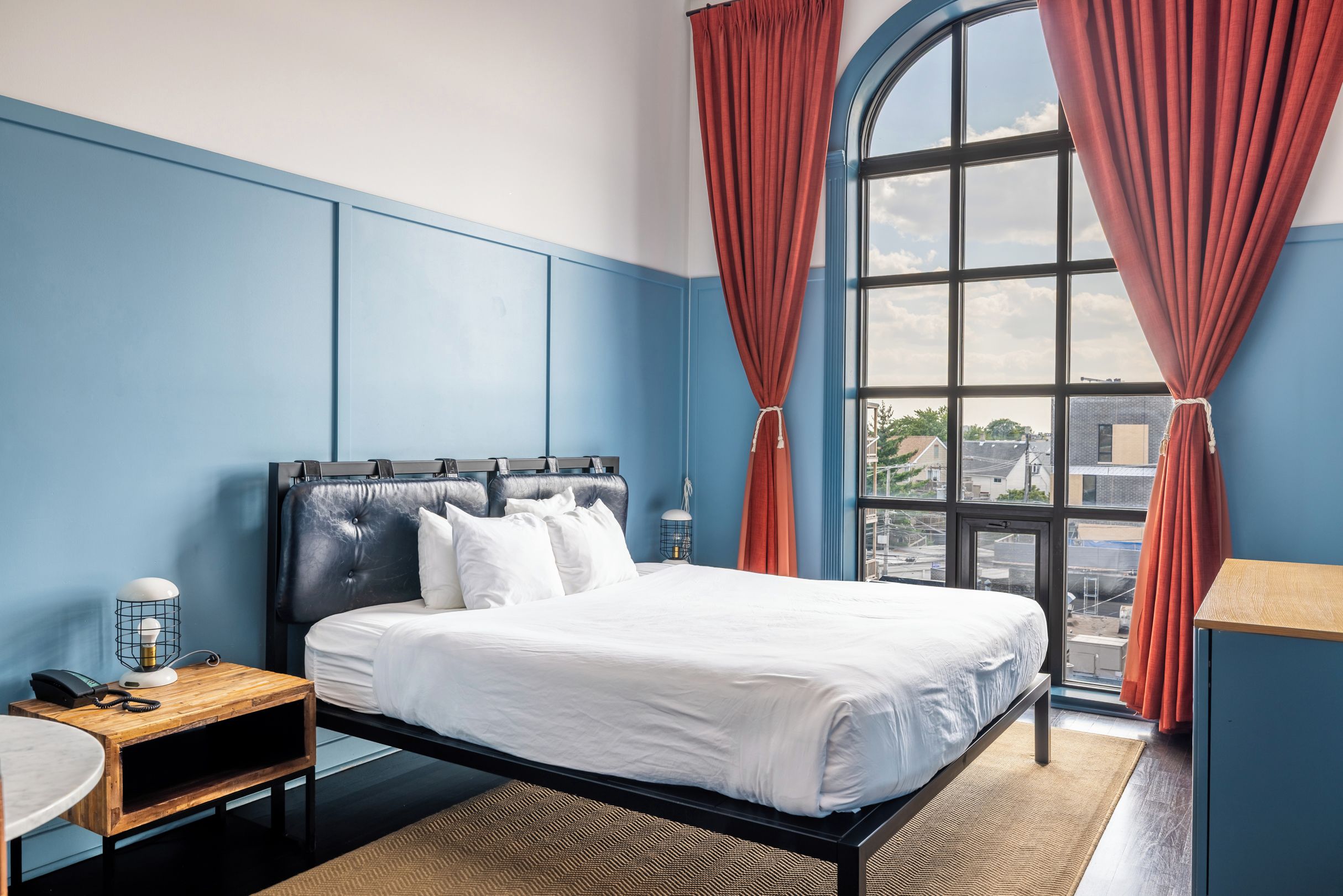 A hotel room with a large bed, a desk, a chair, a lamp, and a view of the city through a large window.