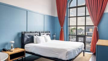 A hotel room with a large bed, a desk, a chair, a lamp, and a view of the city through a large window.