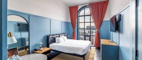 A hotel room with a bed, a desk, a chair, a round table, a mirror, a TV, and a window with curtains.