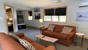 A living room with a brown sofa set, a wooden coffee table, and a small side table. There is a window with blinds, a lamp on the side table, and a framed picture on the wall. - Tahquamenon Suites Lodging (Paradise)