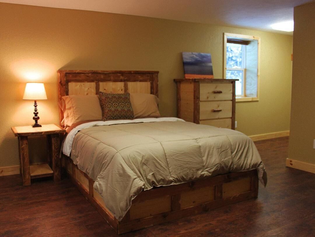 A bedroom with a wooden bed, a nightstand with a lamp, a dresser, and a window with a view.