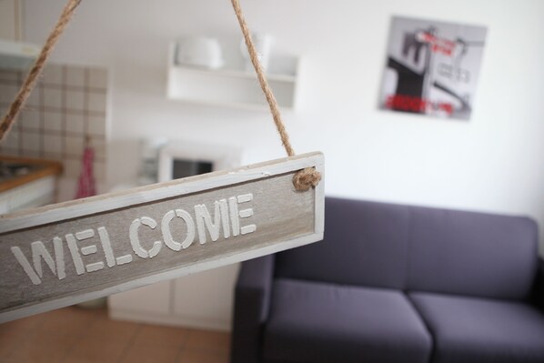 A wooden welcome sign hanging from a rope in a modern living room with a gray sofa and a white cabinet in the background.