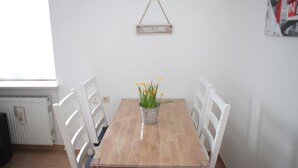 Une table à manger avec des chaises blanches, une pancarte d’accueil suspendue et une plante en pot ornée de fleurs jaunes.