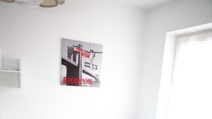 Un lit simple avec une couvre-lit à motifs floraux, un mur blanc orné d’un tableau encadré et une fenêtre avec des stores.