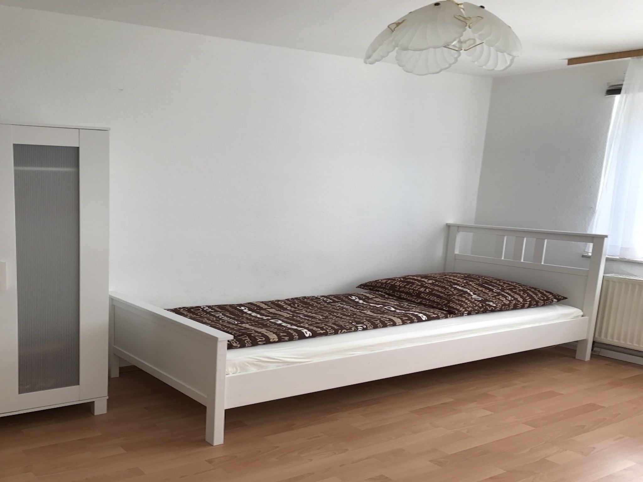 Ein weißes Bett mit gemusterten Betttextilien, ein weißer Kleiderschrank und ein weißer Deckenventilator in einem Raum mit Holzboden.