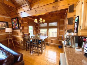 Un interior acogedor de cabaña con una mesa de comedor de madera, sillas, un sofá y una zona de cocina con cafetera.