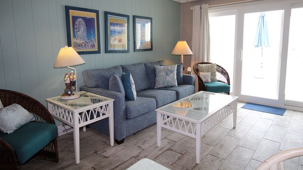 A room with a blue sofa, two white side tables with lamps, wicker chairs, and a glass coffee table. - 13 on the beach 10, Ocean City, MD, USA - Beautiful direct ocean front 3 bedroom 2 bath townhome (Ocean City)