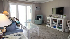 A living room with a sofa, a glass coffee table, a teal armchair, a TV on a cabinet, and a fan. - 13 on the beach 10, Ocean City, MD, USA - Beautiful direct ocean front 3 bedroom 2 bath townhome (Ocean City)