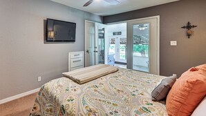 Un dormitorio con cama, ventilador de techo, televisión, puerta corrediza de vidrio y un pequeño armario blanco.