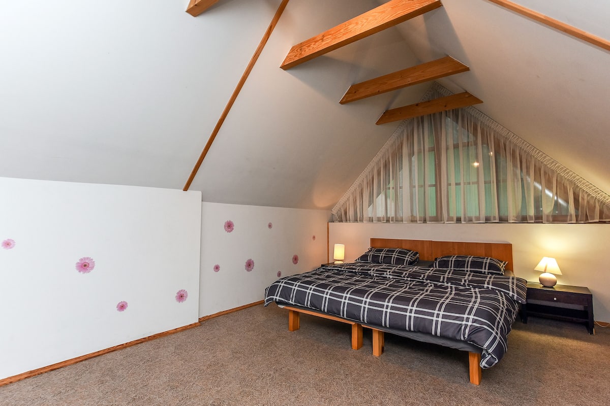 Un dormitorio con techo inclinado, una cama con colcha a cuadros blanco y negro, una mesita de noche con lámpara, y una ventana con cortinas traslúcidas.