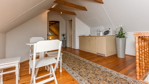 A cozy attic bedroom with a wooden ceiling, a dining area with white furniture, and a kitchen with a wooden cabinet and a large potted plant.