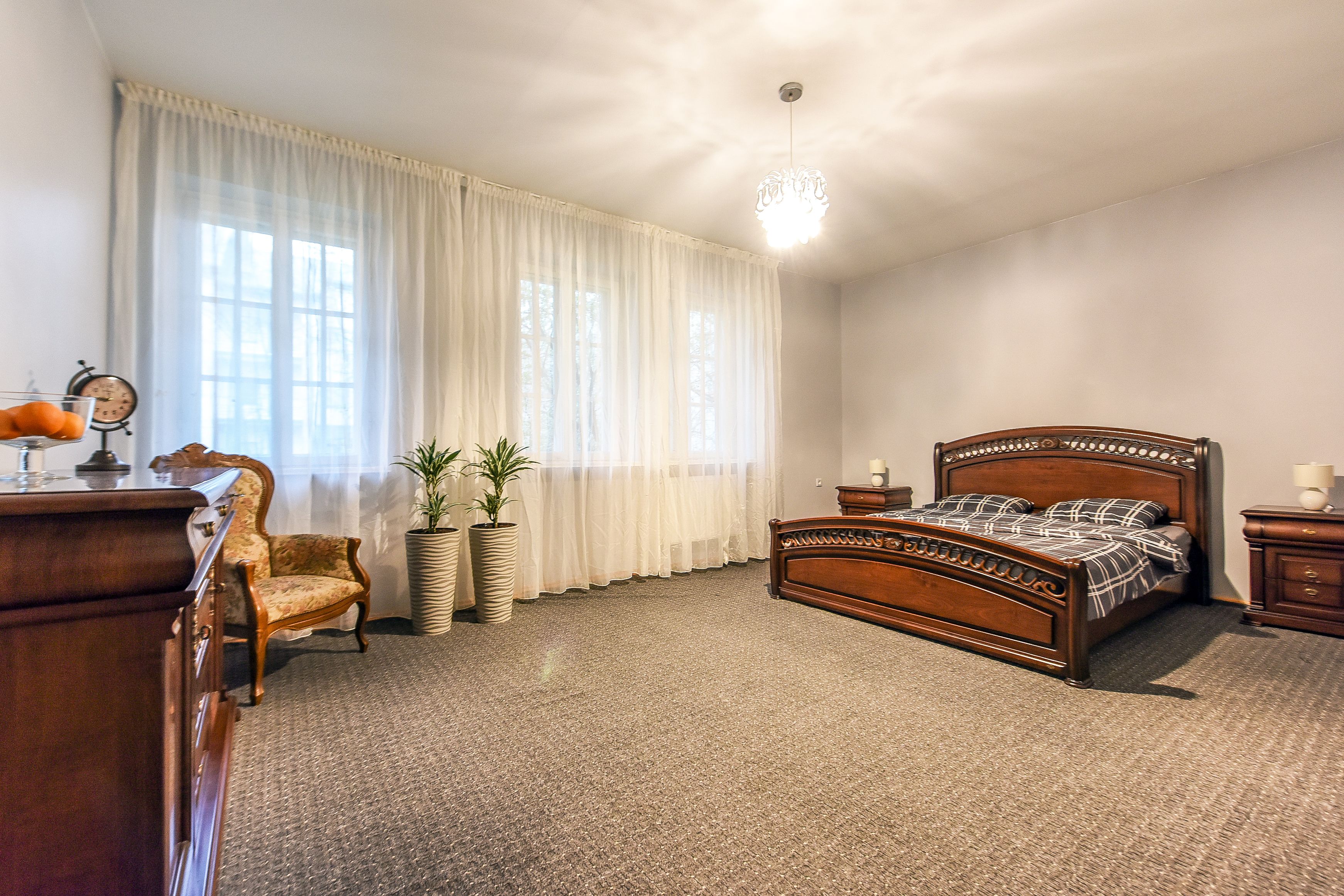 Ein Schlafzimmer mit einem hölzernen Bett, einem Sessel, einem Schminktisch und einem großen Fenster mit durchsichtigen Vorhängen.