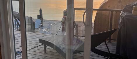 A view from a room with a large glass door overlooking a wooden deck with a grill and outdoor furniture.